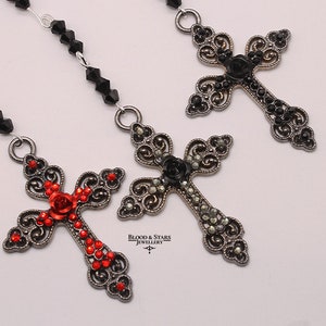 May include: Three ornate silver cross pendants with black and red crystal embellishments. The crosses feature a rose design and are strung on a black beaded chain.