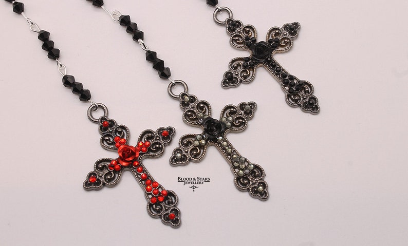 May include: Three ornate silver cross pendants with black and red crystal embellishments. The crosses feature a rose design and are strung on a black beaded chain.