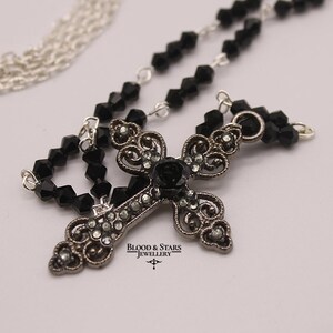 May include: A silver chain necklace with a black crystal bead chain and a silver cross pendant with a black rose and rhinestones. The pendant has the text "Blood & Stars Jewellery" on it.