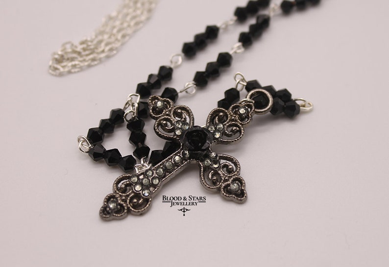 May include: A silver chain necklace with a black crystal bead chain and a silver cross pendant with a black rose and rhinestones. The pendant has the text "Blood & Stars Jewellery" on it.