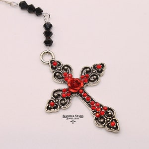 May include: A silver filigree cross pendant with red rhinestone accents and a red rose in the center. The pendant is attached to a black beaded chain.
