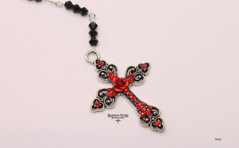 May include: A silver filigree cross pendant with red rhinestone accents and a red rose in the center. The pendant is attached to a black beaded chain.