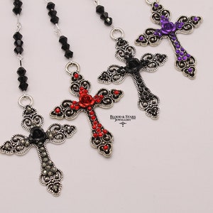 May include: Three silver cross necklaces with black, red, and purple jeweled accents. Each cross features a jeweled rose at the center. The necklaces are on a white background. "Blood & Stars Jewellery" is printed below the crosses.