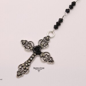 May include: A silver cross pendant with black diamond accents and a black rose in the center. The cross is attached to a black beaded chain.