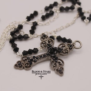 May include: A silver chain necklace with a black crystal cross pendant. The cross is adorned with black crystals and a black rose in the center. The pendant is attached to a silver chain with a silver clasp. The necklace is labeled "Blood & Stars Jewellery".