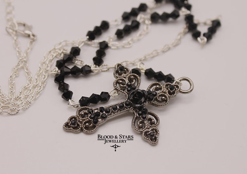 May include: A silver chain necklace with a black crystal cross pendant. The cross is adorned with black crystals and a black rose in the center. The pendant is attached to a silver chain with a silver clasp. The necklace is labeled "Blood & Stars Jewellery".