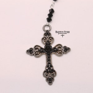 May include: A silver cross pendant with black rhinestone accents. The cross is ornate and features a delicate filigree design. The pendant is attached to a black beaded chain.