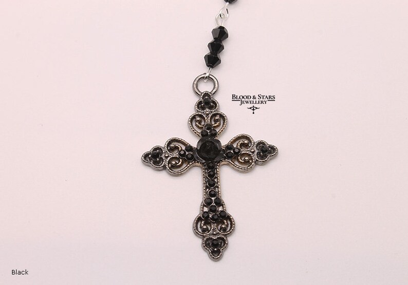 May include: A silver cross pendant with black rhinestone accents. The cross is ornate and features a delicate filigree design. The pendant is attached to a black beaded chain.