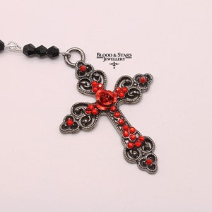 May include: A silver-toned cross pendant with red rhinestone accents and a red rose design. The cross is attached to a black beaded chain.