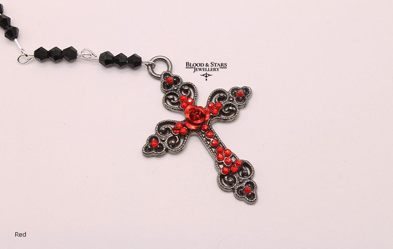 May include: A silver-toned cross pendant with red rhinestone accents and a red rose design. The cross is attached to a black beaded chain.