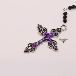 May include: A silver cross pendant with a purple rose and purple rhinestones. The cross is attached to a black beaded chain. "Blood & Stars Jewellery" is printed on the pendant.