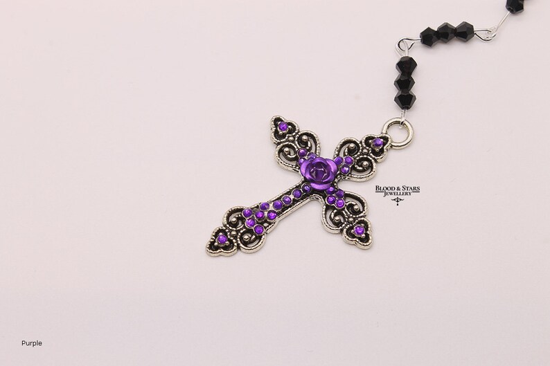 May include: A silver cross pendant with a purple rose and purple rhinestones. The cross is attached to a black beaded chain. "Blood & Stars Jewellery" is printed on the pendant.