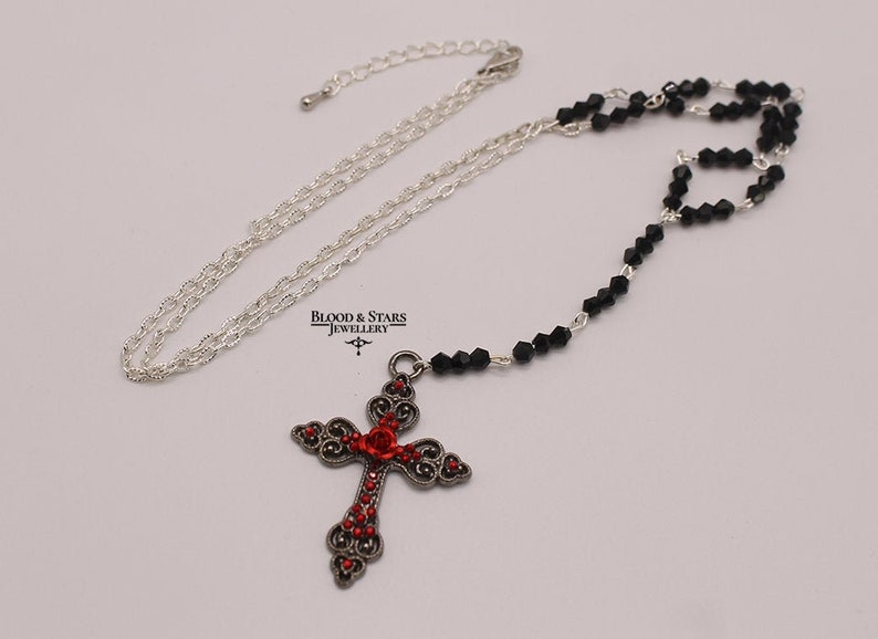 May include: A silver chain necklace with a black bead and red rhinestone cross pendant. The cross has a floral design and the text "Blood & Stars Jewellery" is visible in the image.