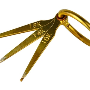 May include: A set of three gold-colored metal jewelry gauges attached to a ring. Each gauge has a pointed tip and is labeled with a karat marking: 18K, 14K, and 10K. The ring is also gold-colored.
