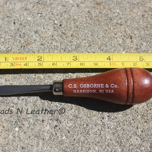 May include: A leatherworking tool with a wooden handle and a metal blade, next to a yellow measuring tape. The tool is marked with "C.S. OSBORNE & Co. HARRISON, NJ USA". The tape measure shows approximately 7 inches.