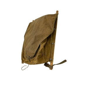May include: A vintage canvas and wood frame backpack with a flap closure and adjustable straps. The backpack is a dark olive green color and has a worn look.