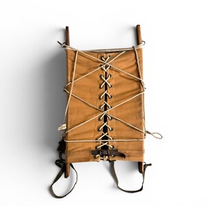 May include: A tan canvas pack frame featuring a central lace-up closure with white cord, secured by metal grommets. The structure includes two vertical wooden side rails, a dark brown leather strap at the base, and adjustable olive green webbing straps.