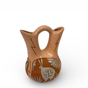 May include: A tan-colored pottery vase with a distinctive handle and two openings. The vase features etched designs, including lightning bolts and a stylized animal figure with silver accents. The overall design is symmetrical and the vase has a rounded base.