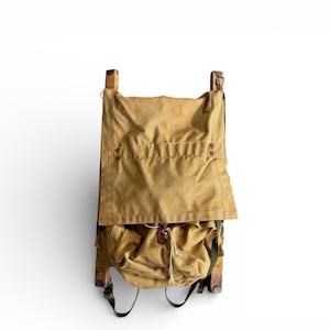 May include: A vintage backpack with a wooden frame and tan canvas. The top flap is secured with small buttons, and the bottom section has a drawstring closure. Black straps and a small pocket are visible.