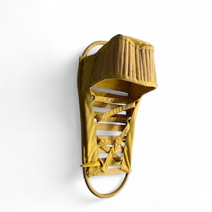 May include: A vintage snowshoe with a yellow fabric and wooden frame. The snowshoe features a laced design and a curved wooden handle. The top section has a woven bamboo-like design. The overall aesthetic is rustic and handcrafted.