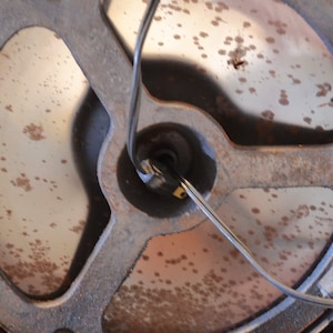 May include: Close-up of the underside of a round, metal lamp base with a black wire attached. The base is rusty and has a circular opening in the center.