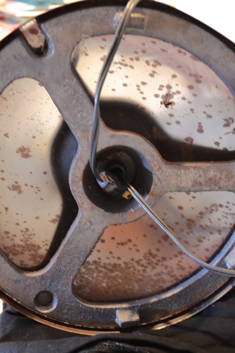 May include: Close-up of the underside of a round, metal lamp base with a black wire attached. The base is rusty and has a circular opening in the center.