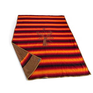 May include: A rectangular blanket with horizontal stripes in shades of red, orange, and black. The blanket has a brown underside and a deer graphic in the center. The blanket is made of a soft material.
