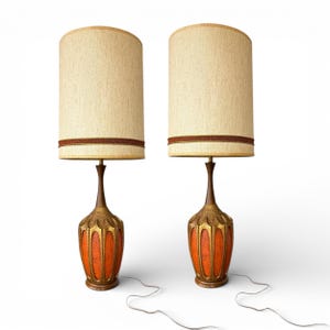 May include: Two vintage table lamps with beige fabric shades and ornate bases. The bases are a combination of orange, gold, and brown, with a decorative pattern. The lamps have a mid-century modern aesthetic and are set against a white background.