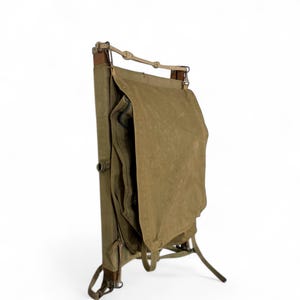 May include: An olive green canvas backpack with a wooden frame. The backpack has a large flap and multiple pockets. The frame is made of wood and leather straps. The backpack is designed to stand upright.