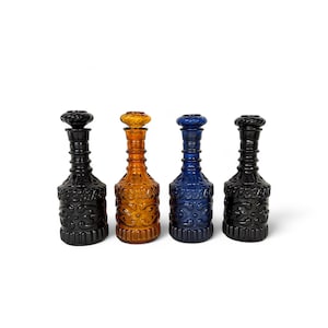 May include: Four decorative glass bottles with stoppers. The bottles are in shades of black, amber, and blue. Each bottle has a textured, patterned design and a matching stopper. The bottles are arranged in a row against a white background.