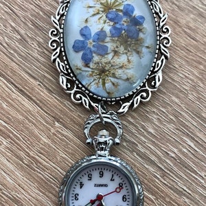 May include: A silver locket with a delicate floral design and a small pocket watch attached. The locket features a pressed flower design with blue flowers and white blossoms. The pocket watch has a white face with black numbers and a red second hand. The watch face reads "QUARTZ".