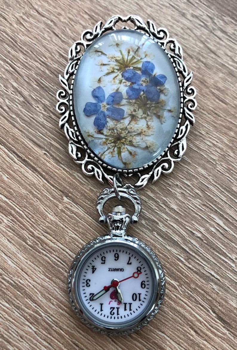 May include: A silver locket with a delicate floral design and a small pocket watch attached. The locket features a pressed flower design with blue flowers and white blossoms. The pocket watch has a white face with black numbers and a red second hand. The watch face reads "QUARTZ".