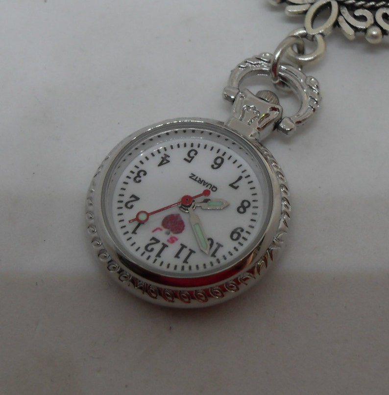 May include: A silver pocket watch with a white face and black numbers. The watch has a small red heart with the words "I love you" on it. The watch is on a white background.