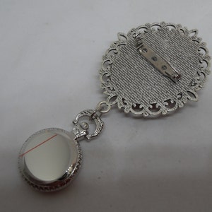 May include: Silver tone pocket watch pendant with a decorative filigree design. The watch has a round face and a silver tone chain attached to a decorative filigree brooch.