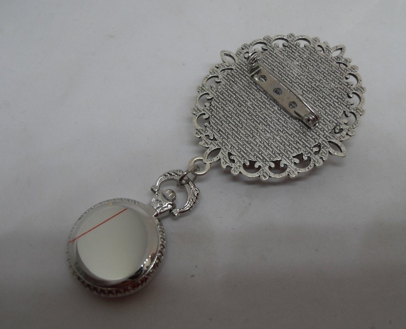 May include: Silver tone pocket watch pendant with a decorative filigree design. The watch has a round face and a silver tone chain attached to a decorative filigree brooch.