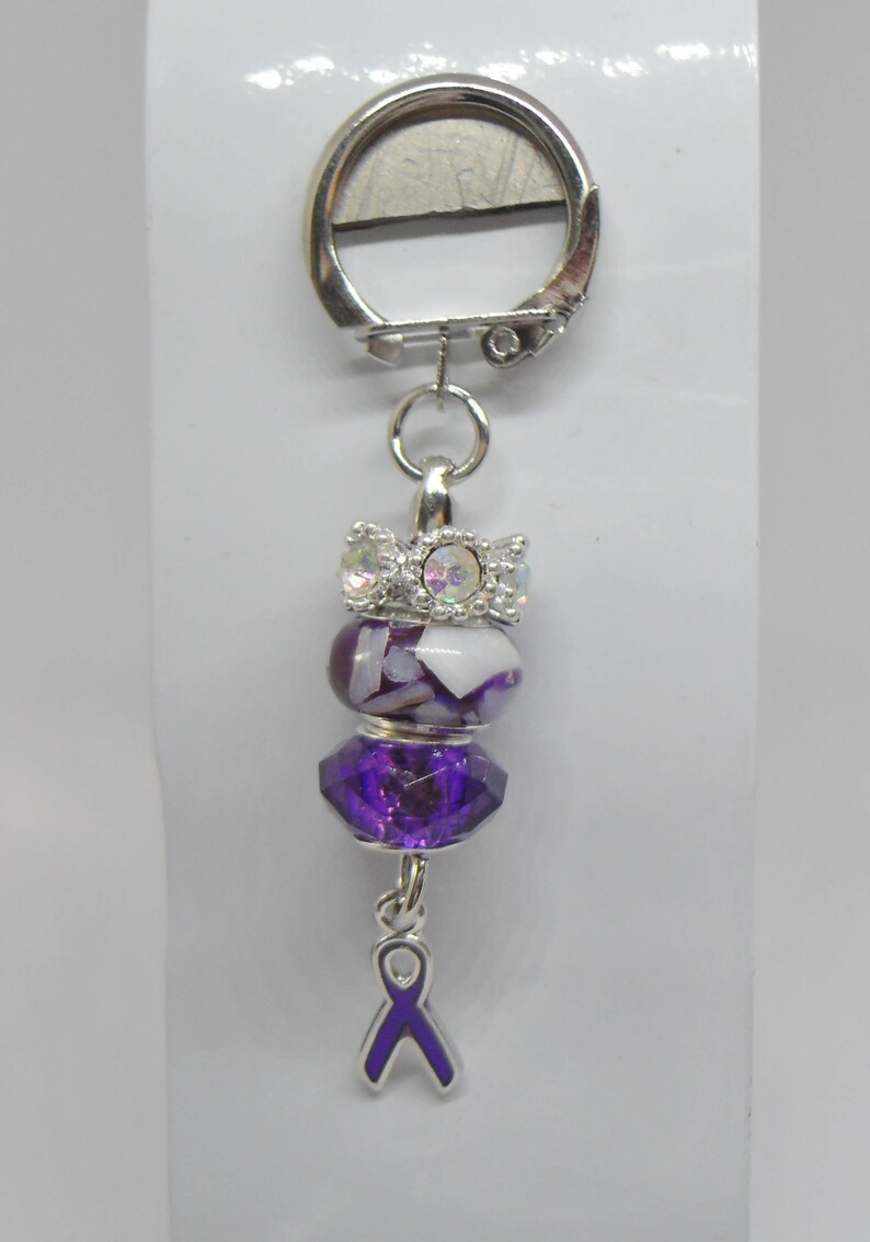 Puede incluir: Un llavero plateado con un dije de cinta morada, una cuenta con patr&oacute;n morado y blanco y una cuenta de cristal transparente con detalles plateados.