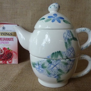 May include: A white ceramic teapot and teacup set with a blue and green floral design. The teapot has a lid and a spout. The teacup has a handle. The set is on a brown hessian background. The box of tea in the image says "Twinings Pomegranate & Raspberry 20 tea bags".