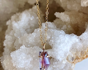 May include: A gold-colored chain necklace with a pendant featuring a faceted, multi-colored gemstone. The gemstone displays shades of purple, pink, and brown. The necklace is displayed against a white, textured background, highlighting the jewelry's details.