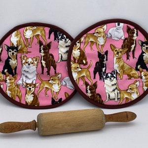May include: Two round pink pot holders with a brown trim, patterned with various Chihuahua dog illustrations. A wooden rolling pin rests below the pot holders. The pot holders are designed for kitchen use.