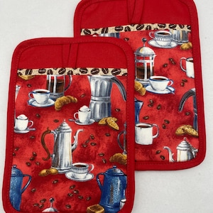 May include: Two red oven mitts with a coffee-themed pattern. The pattern includes coffee pots, cups, and coffee beans on a red background. The top of the mitts has a patterned band with coffee beans. The mitts are designed for kitchen use.