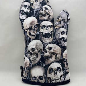 May include: Black oven mitt with a pattern of white skulls on a black background. The skulls are arranged in a random pattern and are all facing forward.