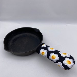 May include: Black cast iron skillet with a black and white patterned oven mitt. The oven mitt has a design of white eggs on a black background.
