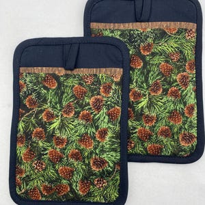 May include: Two navy blue pot holders featuring a pine cone and evergreen needle pattern. They have a dark brown trim and a hanging loop. The background is white.