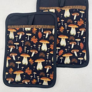 May include: Two black oven mitts with a brown trim and a mushroom and leaf pattern.