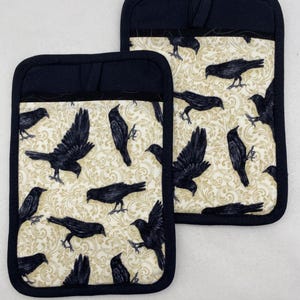 May include: Two black pot holders with a black crow pattern on a cream background. The pot holders have a black border and a loop for hanging. The crows are in various poses, some with wings spread.