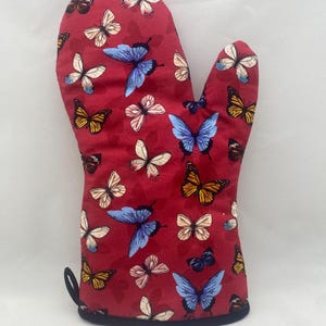 May include: Red oven mitt with a black trim and loop. The mitt is covered in a pattern of butterflies in shades of blue, yellow, white, and black. The background is a solid red.