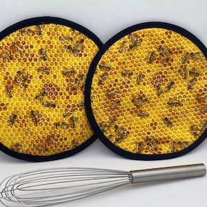 May include: Two round, yellow and black pot holders with a honeycomb and bee pattern. The pot holders have a black border. A stainless steel whisk is in the foreground.
