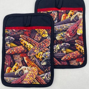 May include: Two black pot holders with a vibrant print of colourful Indian corn. Each pot holder has a red stripe and a loop for hanging. The corn features shades of red, yellow, orange, and purple.