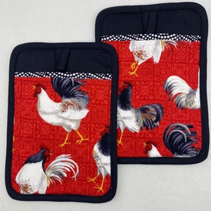 May include: Two square pot holders with a red background featuring white and black roosters. The pot holders have a black border and a black loop for hanging. The top of the pot holders have a black band with white polka dots.