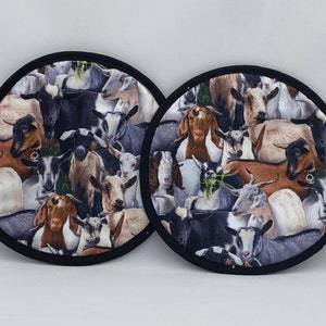 May include: Two round pot holders with a black trim. The fabric features a collage of goats in various colors, including brown, black, and white. The pot holders are designed to protect surfaces from heat.